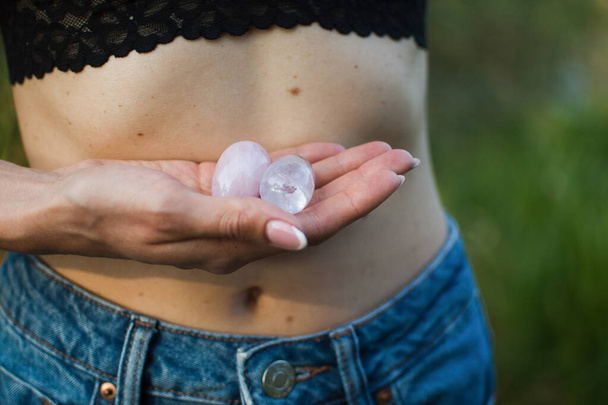 Weibliche Hände, die zwei Yoni-Eier für Vumfit, Imbuilding oder Meditation halten, bestehen aus rosa Quarz und transparentem violettem Amethyst über ihrem nackten Bauch im Freien. - Foto, Bild