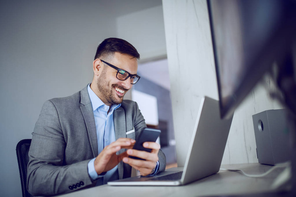 Cheerful attractive classy caucasian unshaven businessman in suit and with eyeglasses sitting in office, using smart phone and looking at laptop. - Foto, afbeelding