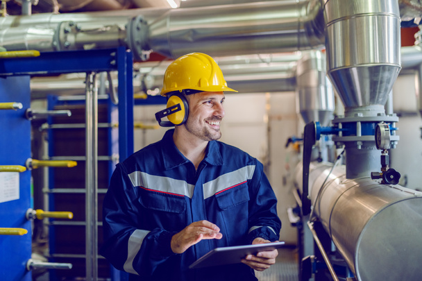 Dedicated smiling factory worker standing next to boiler and holding tablet. Worker is dressed in protective uniform, having hardhat and antiphons. - Foto, imagen