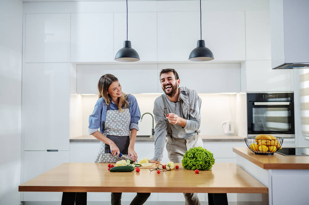 Young charming smiling caucasian woman in apron standing in kitchen and cutting cucumber while talking with her boyfriend. Man holding cherry tomato and talking about healthy lifestyle. - Фото, зображення