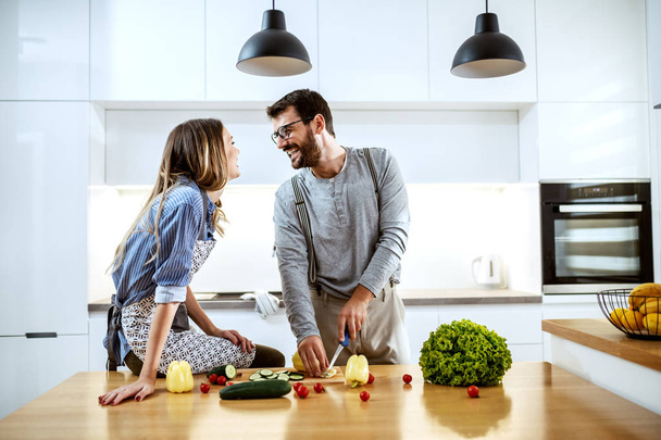 Young attractive caucasian woman in apron sitting on kitchen counter and talking with her boyfriend. Man looking at woman and cutting pepper. On kitchen counter are all sorts of vegetables. - Photo, Image