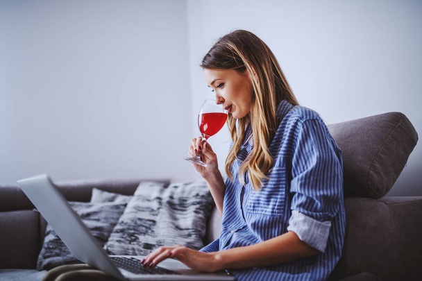 Vista lateral da linda mulher caucasiana em camisa despojada sentada no sofá na sala de estar, bebendo vinho e usando laptop
. - Foto, Imagem