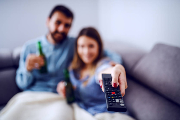 リビングルームにある毛布で覆われた魅力的な白人のカップルは、テレビを見てビールを飲んでいます。リモコンを持ってチャンネルを変える女。リモートコントロールに焦点を当てる. - 写真・画像
