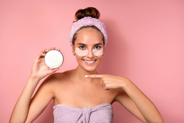 A beautiful girl with a bandage on head with patches under eyes stands on a pink background, points finger at the cream in hand - Fotografie, Obrázek
