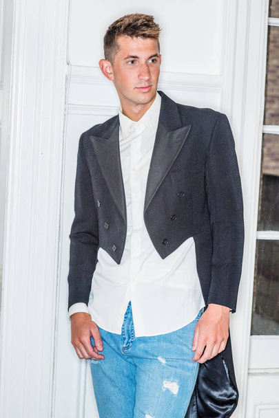 Portret młodego przystojniaka w domu w Nowym Jorku. 20-letni mężczyzna w czarnej jaskółce - płaszcz ogonowy, biała koszula, niebieskie dżinsy rozdarte dżinsy, stojący przy białych drewnianych drzwiach z oknem - Zdjęcie, obraz