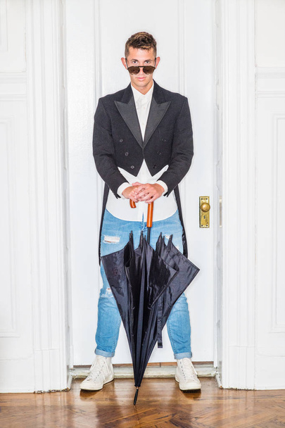 Jonge man mode in New York City. 20 jaar oud man draagt zwarte Swallow - Tailed jas, wit shirt, blauw gescheurde denim jeans, sneakers, zonnebril, met zwarte stok paraplu met haak handvat, - Foto, afbeelding