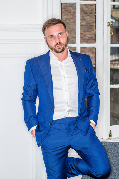 Portrait of Young Man at home in New York City. Young guy with beard, wearing blue suit, unbuttoned, white shirt, hands on pocket, standing against white wooden door with window, looking at you. - Fotoğraf, Görsel