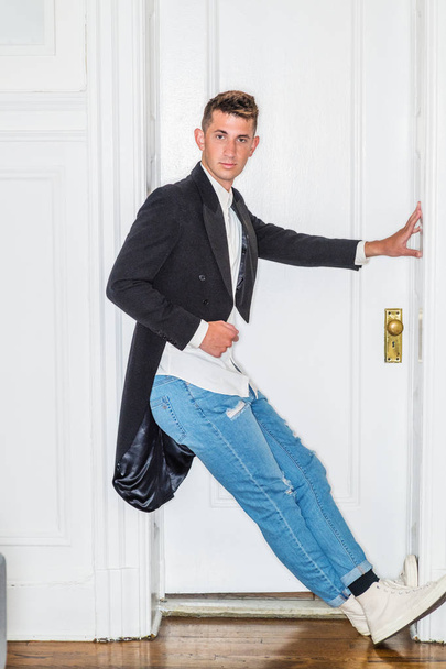 Portrait of Young Handsome Man in New York City. 20 years old man wearing black Swallow - Tailed coat, white shirt, blue ripped Denim jeans, sneakers, standing by white wooden doorway, having fun - Fotografie, Obrázek