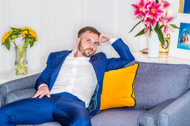 junger müder Mann mit Bart, entspannt zu Hause in New York City, trägt blauen Anzug, weißes Hemd, sitzt auf einem Sofa mit gelbem Wurfkissen, die Hand berührt den Kopf, denkt. Blumen auf Hintergrund - Foto, Bild