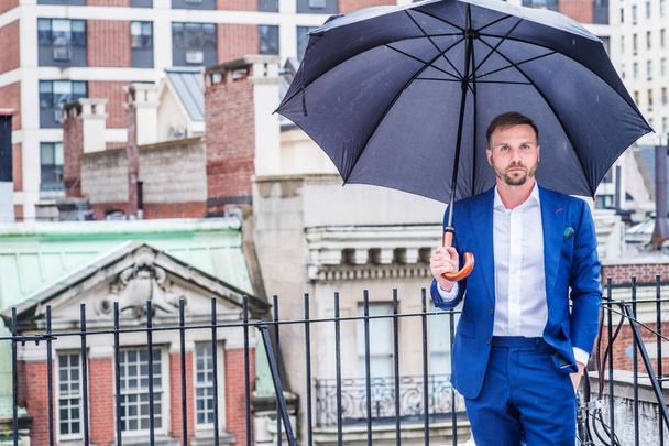 Little raining, drizzle, wet feel. Young man with beard, relaxing, thinking outside in raining day, wearing blue suit, white shirt, holding black umbrella, standing on roof top in New York City - Photo, Image