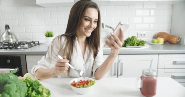 Woman Eating Salad in Kitchen Holding Smartphone - Materiaali, video