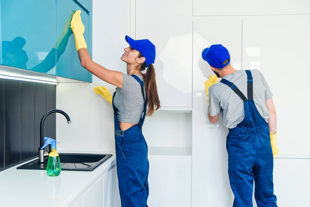 Couple of professional cleaners in blue uniform cleaning the furniture with wipes and spray in the contemporary kitchen. - Фото, изображение