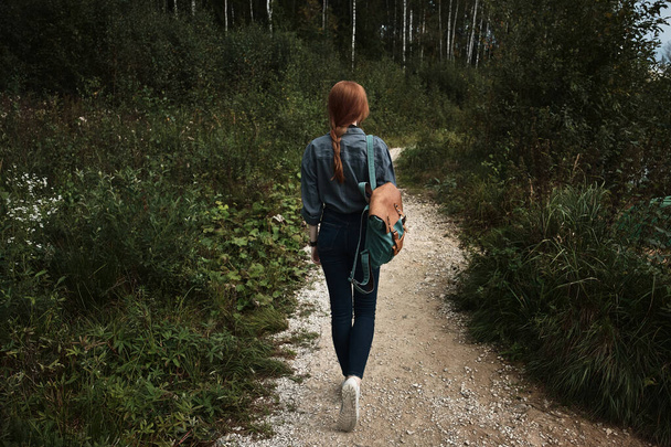 A slender girl with a red braid in a blue shirt with a backpack is walking along a forest path, a frame from the back. - Foto, imagen