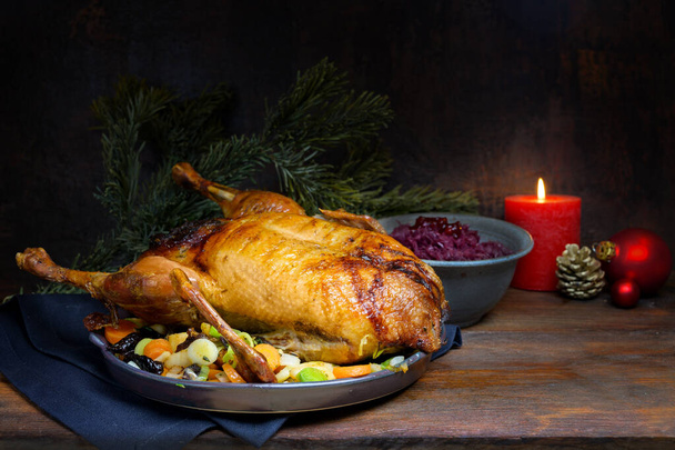 Pečená kachna jako vánoční menu s červeným zelím a zeleninou, slavnostní jídlo na venkovském dřevěném stole s hořící svíčkou a dekorací, tmavě hnědé pozadí s kopírovacím prostorem - Fotografie, Obrázek