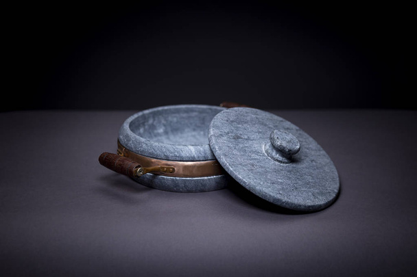 Moody image of a heavy marble stone casserole  with brass element and wooden handle with the lid half next to it on a smooth grey seamless surface - Fotoğraf, Görsel