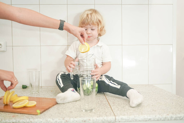 Портрет чарівної матері і маленької дитини, що готує овочевий напій з водою на білій кухні
 - Фото, зображення