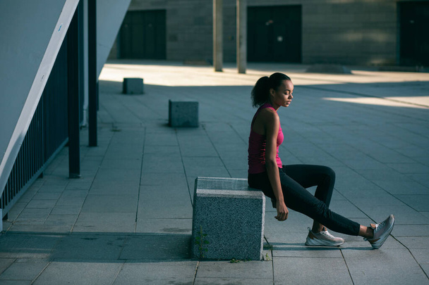 Calm beautiful sportswoman sitting outdoors on the stone cube and looking at her foot - Fotoğraf, Görsel