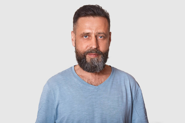 Studio shot of middle aged man with beard dresses grey casual t shirt, posing isolated over light background, attractive male looking directly at camera with calm facial expression. People concept. - Foto, Imagem