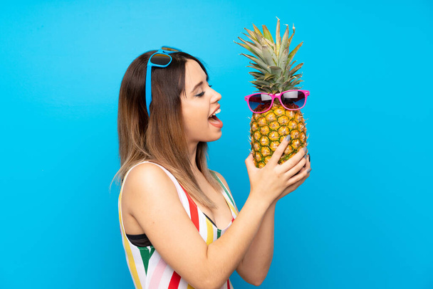 Young woman in summer holidays over blue background holding a pineapple with sunglasses - Photo, image