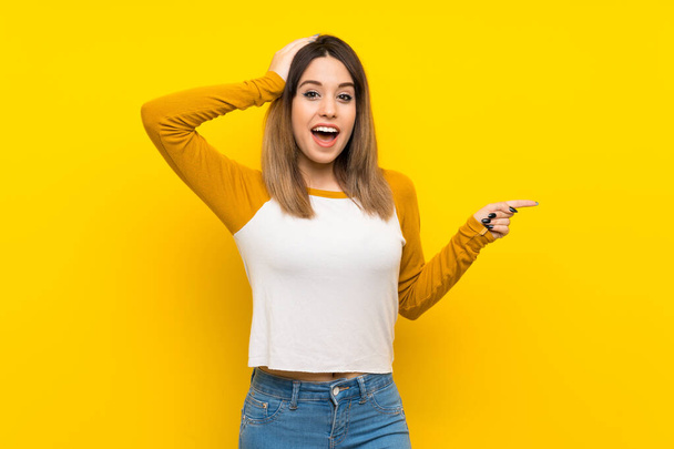 Красивая молодая женщина над изолированной желтой стеной удивлена и указывая пальцем в сторону
 - Фото, изображение