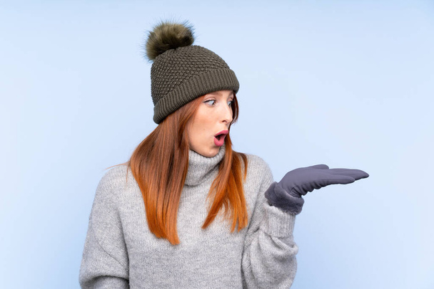 Young redhead Russian woman with winter hat over isolated blue background holding copyspace imaginary on the palm - Photo, Image