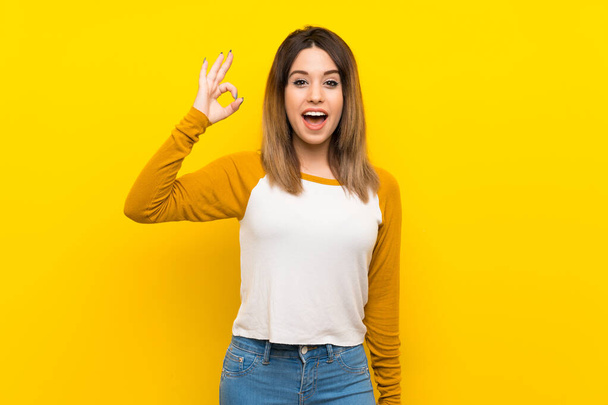 Гарненька молода жінка над ізольованою жовтою стіною здивувала і показала знак ОК
 - Фото, зображення