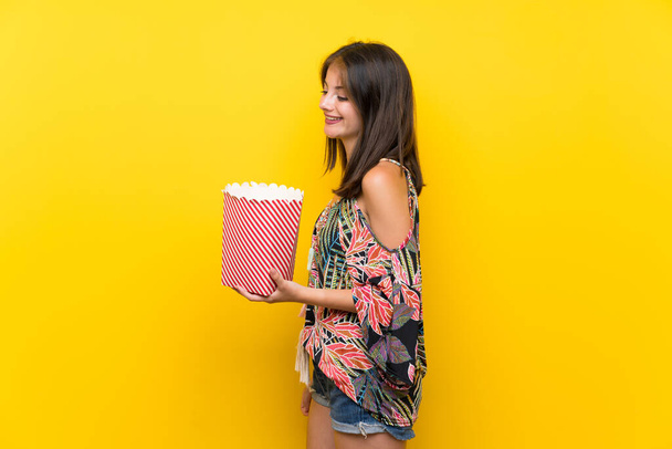 Caucasian girl in colorful dress over isolated yellow background holding a bowl of popcorns - Fotoğraf, Görsel