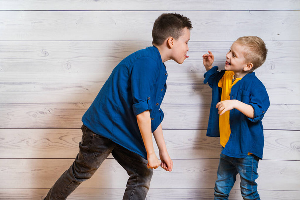 Two boys in casual clothes in white wooden background showing tongues to each other. Two brothers playing. Elder and smaller brothers. Happy childhood concept. Frienship. - Foto, Imagen