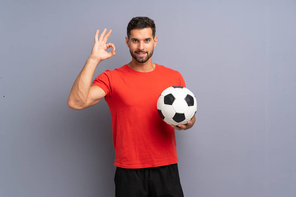 Fešák mladý fotbalista muž přes izolované bílé zdi ukazující ok znamení s prsty - Fotografie, Obrázek
