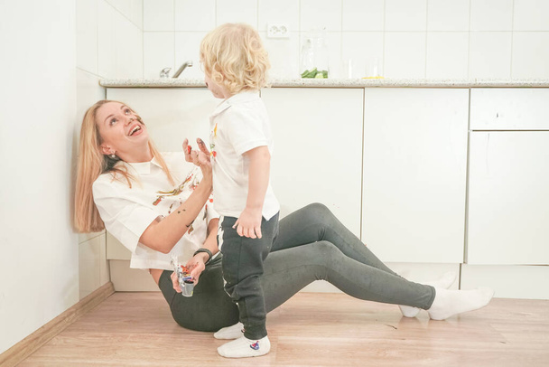 Neşeli bir anne ve neşeli bir çocuk yere beyaz tişörtlerle oturur ve boyayla birbirlerini boyarlar. - Fotoğraf, Görsel