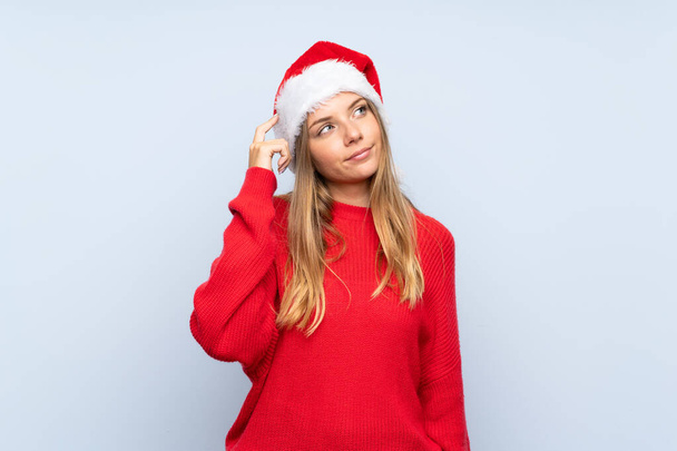 Lány karácsonyi kalap felett elszigetelt kék háttér kétségek és zavaros arckifejezés - Fotó, kép