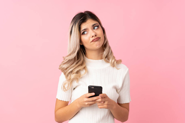 Подросток на изолированном розовом фоне с помощью мобильного телефона с наушниками и думать
 - Фото, изображение