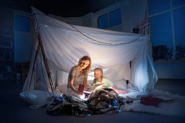 Madre e hija sentadas en un tipi, leyendo cuentos con la linterna
 - Foto, Imagen