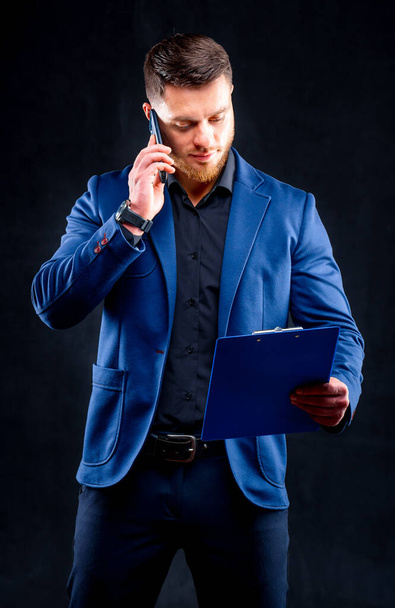 Hombre de negocios de confianza está hablando por teléfono con la carpeta en la mano aislado sobre fondo negro en el estudio
. - Foto, imagen