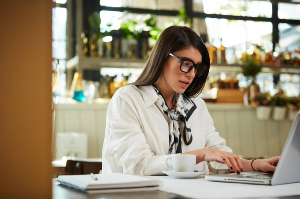 Attractive businesswoman with glasses sitting in cafe and using laptop. On table are laptop, notebook and cup of coffee. Hands are on keyboard. - Photo, Image