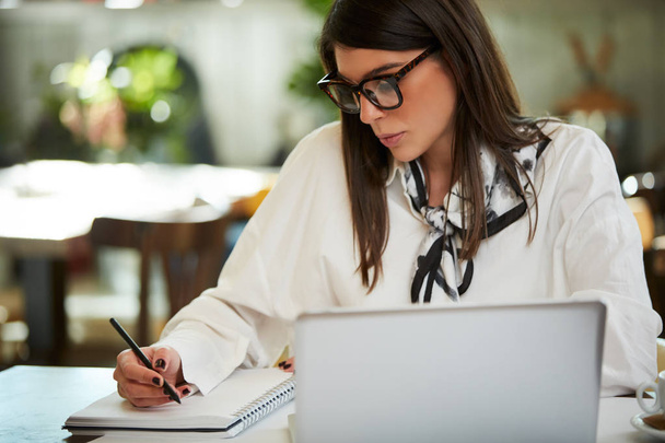 Urocza, biała bizneswoman w okularach siedząca w kawiarni i pisząca zadania w notatniku. Przed nią na stole jest laptop.. - Zdjęcie, obraz