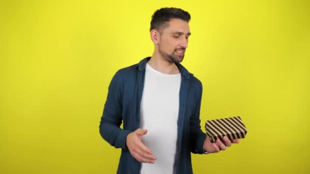 A young man in a white T-shirt throws a box with a gift from hand to hand and smiles, yellow background. Holidays concept: New Year, Christmas, Valentine's Day birthday. 4kfootage - Кадри, відео