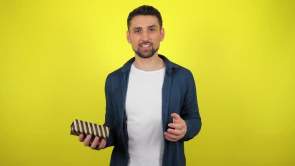 A young man in a white T-shirt throws a box with a gift from hand to hand, looks at the camera and smiles, yellow background. Holidays concept: New Year, Christmas, Valentine's Day birthday. 4kfootage - Materiał filmowy, wideo