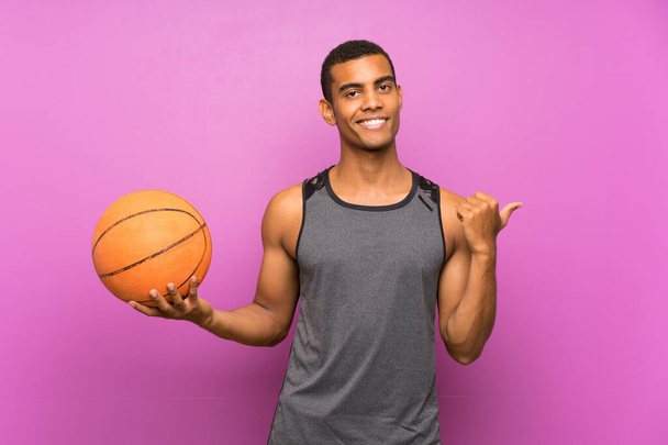 Mladý sportovní muž s míčkem basketbalu přes izolovanou fialovou stěnu ukazující na stranu prezentovat produkt - Fotografie, Obrázek