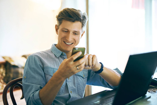 Πορτρέτο ενός ενθουσιασμένου ευτυχισμένος όμορφος άντρας χρησιμοποιώντας το τηλέφωνο και φορητό υπολογιστή στο σπίτι - Φωτογραφία, εικόνα