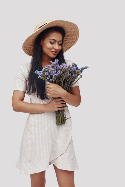attraktive junge Frau mit Hut lächelnd und mit Blumenstrauß vor grauem Hintergrund - Foto, Bild