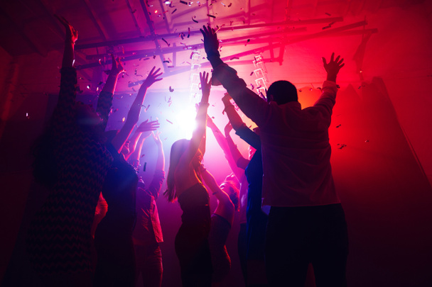 Ένα πλήθος ανθρώπων σε σιλουέτα σηκώνει τα χέρια τους ενάντια στο πολύχρωμο φως νέον στο φόντο κόμμα - Φωτογραφία, εικόνα