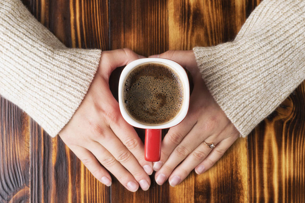 Female hands in a woolen sweater hold a cup of coffee on a wooden table - Фото, зображення