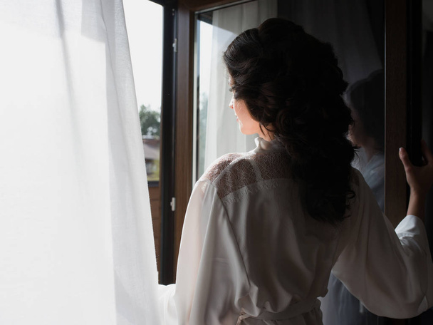  portré gyönyörű vidám barna nő fehér selyem köntösben áll az ablaknál reggel - Fotó, kép