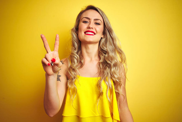 Νεαρή attactive γυναίκα φορώντας t-shirt στέκεται πάνω από κίτρινο απομονωμένο φόντο δείχνει και δείχνει προς τα πάνω με τα δάχτυλα νούμερο δύο, ενώ χαμογελά αυτοπεποίθηση και χαρούμενος. - Φωτογραφία, εικόνα