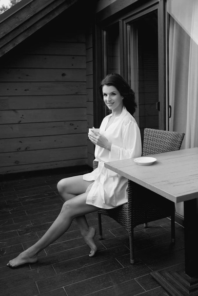 monochroom portret van mooie vrolijke brunette vrouw in witte zijde badjas drinken koffie op balkon - Foto, afbeelding