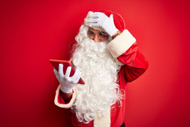 Hombre de mediana edad con traje de Santa Claus usando tableta sobre fondo rojo aislado estresado con la mano en la cabeza, sorprendido con la vergüenza y la cara sorpresa, enojado y frustrado. Miedo y malestar por error
. - Foto, imagen