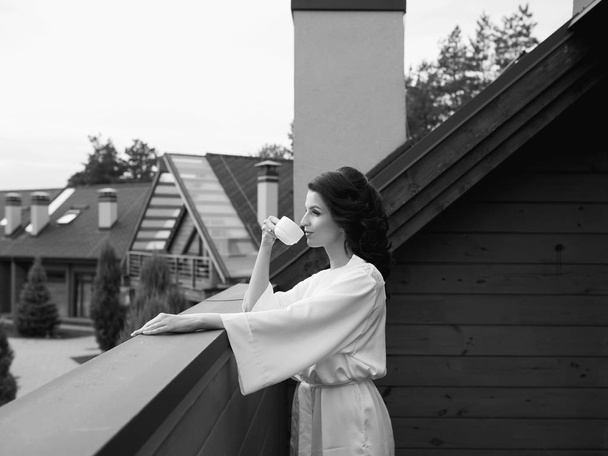 belle femme brune heureuse et joyeuse en robe de soie blanche buvant du café au balcon
 - Photo, image