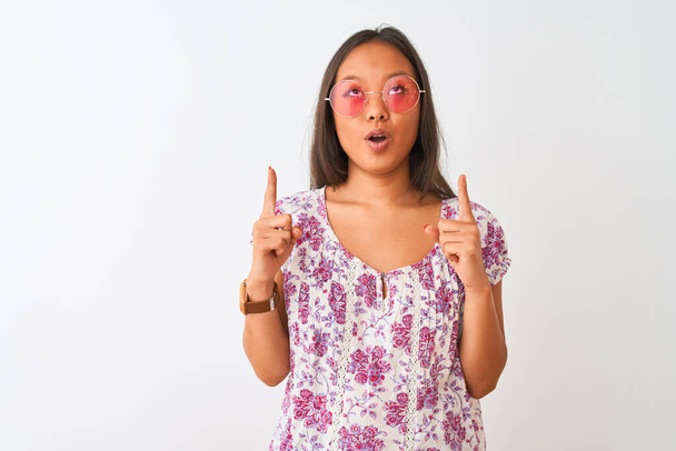 Młoda Chinka w kwiatowej koszulce i różowych okularach na białym tle zaskoczona i zaskoczona patrząc w górę i wskazując palcami i podniesionymi ramionami. - Zdjęcie, obraz