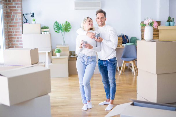 Young beautiful couple standing drinking cup of coffee at new home around cardboard boxes - Photo, Image